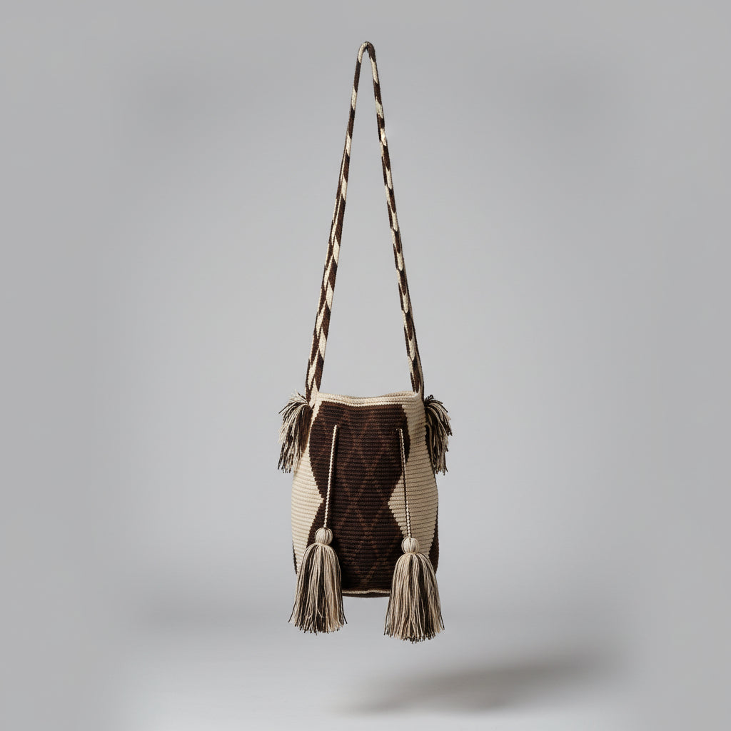 A handwoven Wayuu mochila shoulder bag with a dark brown and beige traditional geometric design pattern and matching cross-body strap, hanging on a white studio wall