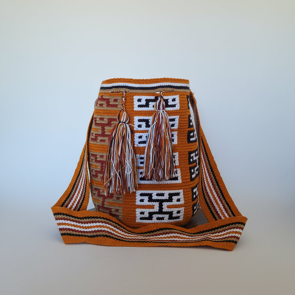 A traditional Wayuu mochila bag with a vibrant ochre body, featuring geometric black and white patterns, two large braided tassels, and a wide striped shoulder strap, displayed in studio against a plain light background.